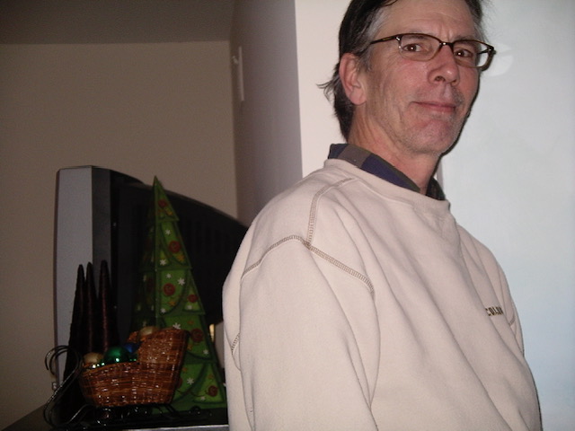 A man in glasses beside a television and a small Christmas tree